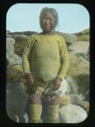 Image of Old Esquimeau woman by stone igloo, holding piece of hide