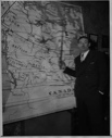 Image of Davis Irwin stands by map showing his planned route to the North Pole by dog sled in 1939 or 1940