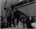 Image of Donald MacMillan seated at the BOWDOIN's wheel. ? standing by, after arriving in Mystic