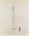 Image of High tower with guy lines during snow storm. Men standing by. Thule AFB. 